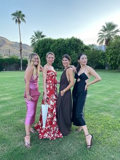 four women in dresses posing for a photo on the grass with palm trees behind them