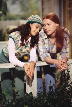 two young women leaning on a rail in front of some bushes and trees, one is touching the other's hand