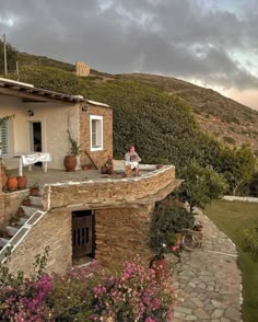 a man sitting on top of a stone building next to a lush green hillside covered in flowers