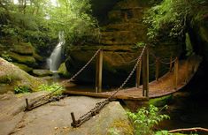 a wooden bridge over a small stream in the woods next to a waterfall with a chain hanging from it's sides