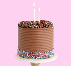 a chocolate birthday cake with sprinkles and two candles on top, against a pink background