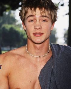 a close up of a shirtless man wearing a chain around his neck with trees in the background