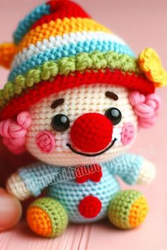 a small crocheted clown doll sitting on top of a pink table next to a person's hand