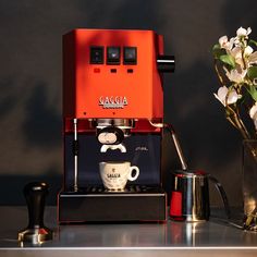 an orange espresso machine sitting on top of a counter next to a vase with flowers