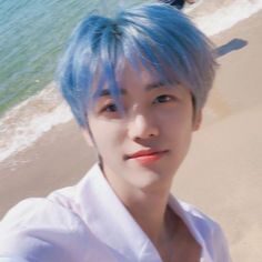 a young man with blue hair standing on a beach next to the ocean and looking at the camera