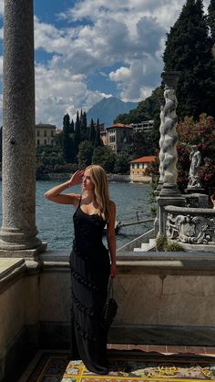 a woman in a long black dress standing on a balcony next to a body of water