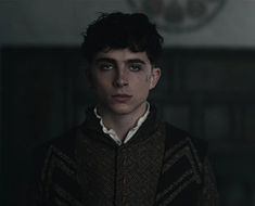 a young man with dark hair wearing a sweater and looking at the camera in a dimly lit room