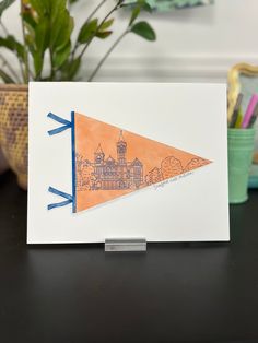 a card with an orange and blue pennant on it sitting next to a potted plant