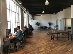 people are sitting at desks in an office with large windows and wooden flooring