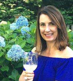 a woman holding a glass of wine in front of blue hydrangea bushes and trees