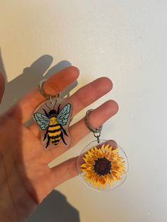 a person's hand holding two key chains with sunflower and bee on them