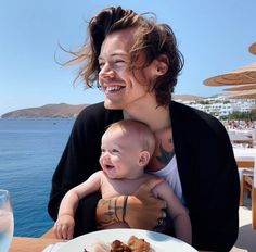 a woman holding a baby while sitting at a table with food and water in front of her