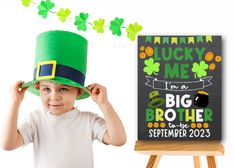 a little boy wearing a green hat and standing next to a sign that says lucky me i'm a big brother