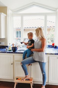 a woman holding a child while sitting on top of a stool in a kitchen next to a window