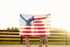 a woman holding an american flag in front of a fence with the sun behind her