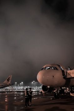 two people standing next to an airplane on the tarmac at night with dark clouds in the background