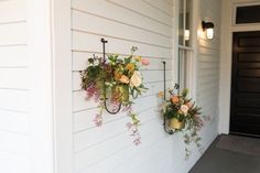 two hanging flower baskets on the side of a white house, one with flowers in it