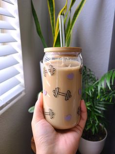 a hand holding a mason jar with a straw in it next to a potted plant