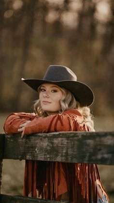 a woman wearing a cowboy hat leaning on a wooden fence with her arms crossed and looking off to the side