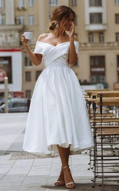 a woman in a white dress is holding a cup and talking on her cell phone