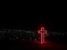 a cross lit up in the dark on top of a hill with city lights behind it