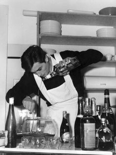 a woman in an apron is pouring something into a glass on the counter next to bottles and glasses