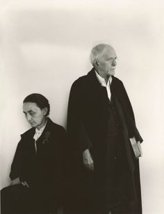 an old man and woman standing next to each other in front of a white wall