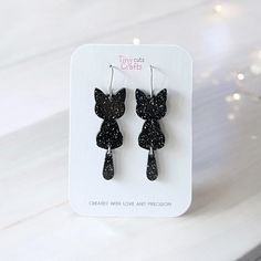 Black cat earrings with glittery details, perfect for Halloween or spooky-themed outfits. These cute and whimsical earrings are crafted from high-quality acrylic with a fun, dangling tail design that adds movement and sparkle. Lightweight and comfortable, they make a great accessory for Halloween parties or everyday spooky style. Ideal for cat lovers or as a festive gift, these black glitter cat earrings bring a touch of charm to any outfit.  Made with stainless steel findings for sensitive ears Halloween Cat Earrings, Party Jewelry With Cat Design And Cat Ears, Party Jewelry With Cat Design, Black Cat Ears Earrings For Party, Cute Cat Design Earrings For Party, Cute Black Party Earrings, Whimsical Earrings, Clay Halloween, Polymer Clay Halloween