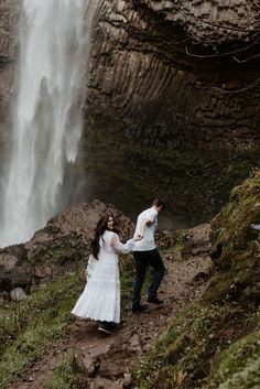 a man and woman holding hands walking up a hill next to a waterfall with water cascading on it