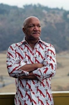 an older man with his arms crossed standing in front of a mountain range and looking off into the distance