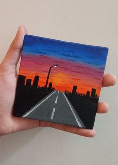 a hand holding up a small painting of a cityscape at sunset with the sun going down