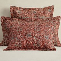 two red pillows sitting next to each other on top of a white bed covered in paisley print