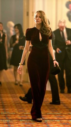 a woman in a long dress walking down a hall with other people behind her and one man wearing a suit