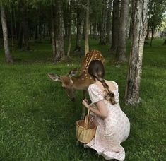 a woman kneeling down in the grass next to a deer that is eating from a basket