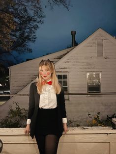 a woman standing in front of a white house wearing a red bow tie and black skirt