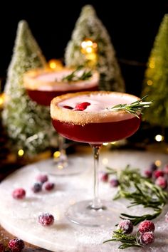 two glasses filled with red liquid and garnished with greenery, surrounded by small christmas trees