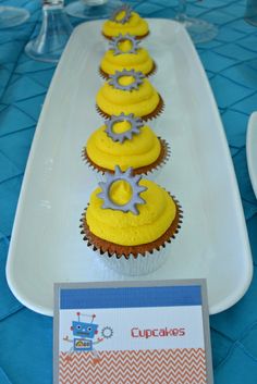 cupcakes with yellow frosting are lined up on a white platter in front of a blue tablecloth