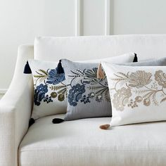 three pillows on a white couch with blue and gold flowers in the middle, along with black tassels