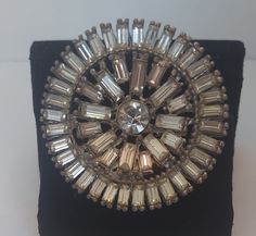 This Art Deco Style Cut Glass Baguette Round Star or Flower Brooch in Silvertone metal is a circular layer of Baguettes encircling 1 larger round Cut Glass Rhinestone. Semi shiny to matte finish silvertone metal encompasses the back with heart shaped openings around the center stone that appear unique.  Measures:   41.65 mm wide around x 3.80 mm thick Silver Rhinestone Round Brooches, Silver Round Brooches With Rhinestones, Silver Round Brooches For Evening, Round Star, Cape May, Star Flower, Art Deco Style, Flower Brooch, Deco Style