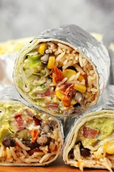 three burritos wrapped in foil on top of a wooden table