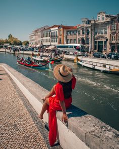 a woman in a red dress and straw hat sitting on a wall looking at boats