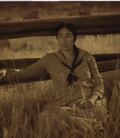 a woman sitting on the ground next to a wooden fence with grass in front of her