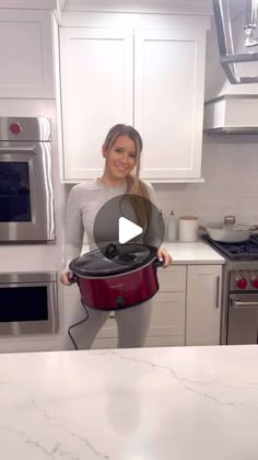 a woman standing in a kitchen holding a red pot with the lid open and smiling