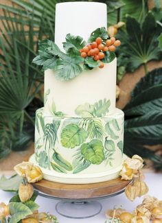 a white and green wedding cake with orange berries on top, surrounded by greenery