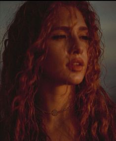 a woman with red curly hair wearing a necklace and looking off into the distance while standing in front of a dark background