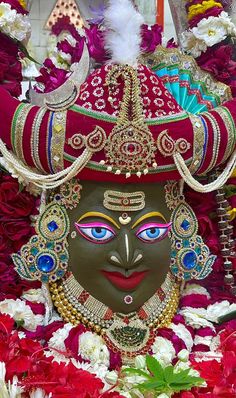 an elaborate mask is displayed in front of red and white flowered garlands with feathers