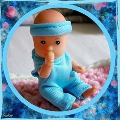 a baby doll wearing a blue outfit and holding a finger up to her mouth while sitting on top of a pink blanket