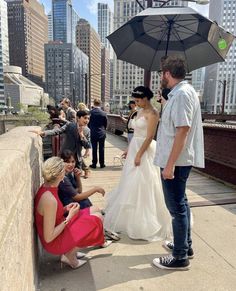 a bride and groom standing under an umbrella on a bridge with other people around them