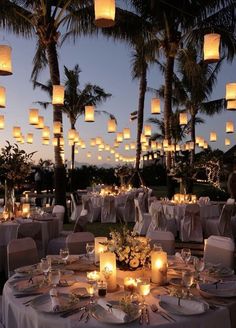 an outdoor dining area with lit candles and paper lanterns in the sky above it, surrounded by palm trees