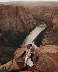 a bride and groom are sitting on the edge of a cliff looking at the river